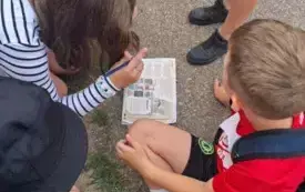 Na asfalcie leży kartka z zagadką, a wok&oacute;ł niej siedzą dzieci, skupione na wsp&oacute;lnym rozwiązywaniu zadania.