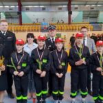 Młodzi zwycięzcy zawodów w kompletnych strojach strażackich. Po bokach stoją przedstawiciele organizatorów wydarzenia. Wszyscy ustawieni w szeregu podczas ceremonii wręczenia nagród.