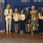 Grupa osób pozuje do zdjęcia podczas uroczystości zakończenia roku sportowego. W centrum znajdują się nagrodzeni zawodnicy trzymający dyplomy, medale lub puchary.
