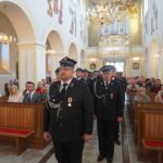 Ludzie siedzą w ławkach w kościele. Po środku stoją druhowie strażacy.