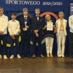 Grupa osób pozuje do zdjęcia podczas uroczystości zakończenia roku sportowego. W centrum znajdują się nagrodzeni zawodnicy trzymający dyplomy, medale lub puchary.