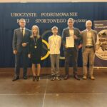 Grupa osób pozuje do zdjęcia podczas uroczystości zakończenia roku sportowego. W centrum znajdują się nagrodzeni zawodnicy trzymający dyplomy, medale lub puchary.