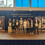 Grupa osób pozuje do zdjęcia podczas uroczystości zakończenia roku sportowego. W centrum znajdują się nagrodzeni zawodnicy trzymający dyplomy, medale lub puchary.