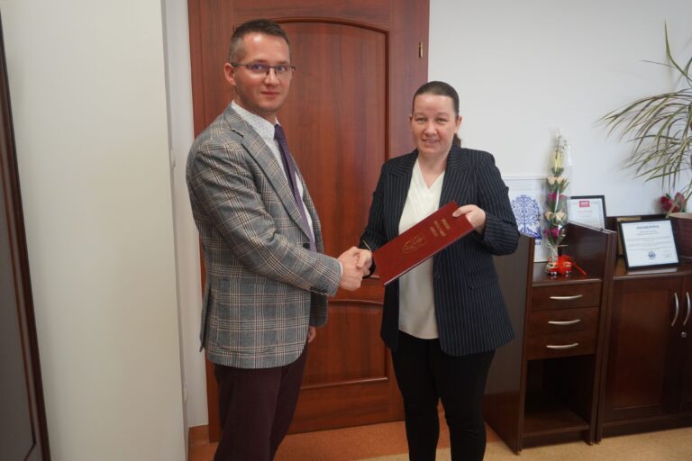 Burmistrz wręcza akt powierzenia stanowiska nowej Pani Dyrektor, która z uśmiechem trzyma w dłoni symboliczną tęczkę.