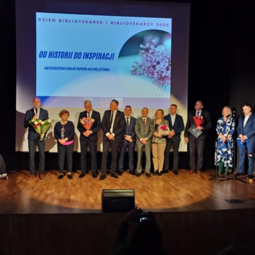 Grupa ludzi stoi na scenie. W tle telebim.