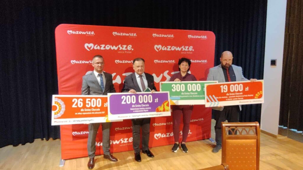 Na zdjęciu widoczni są przedstawiciele Województwa Mazowieckiego oraz gminy Chorzele podczas uroczystego wręczenia dofinansowania. Czterech uczestników uroczystości - trzech mężczyzn i jedna kobieta - pozuje do zdjęcia, trzymając w rękach cztery duże symboliczne czeki. Na każdym z czeków widnieje kwota dofinansowania oraz nazwa zadania, które otrzymało wsparcie finansowe. Osoby ustawione są obok siebie w jednym rzędzie.